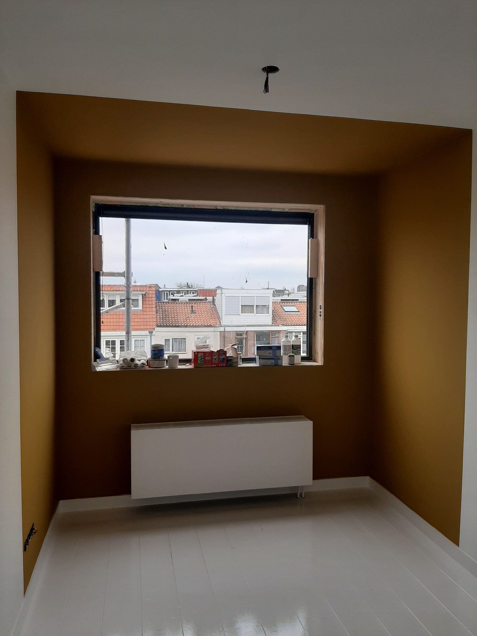Pomieszczenie po malowaniu ścian na kolor musztardowy, z białymi listwami przypodłogowymi i grzejnikiem. Widok z okna na zabudowę miejską.