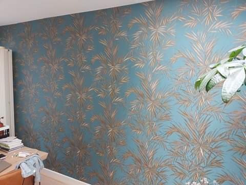Turkusowa ściana z wzorem złotych palm, widok z bliska. Częściowo widoczny blat biurka z książkami i laptopem oraz roślina doniczkowa w prawym górnym rogu.