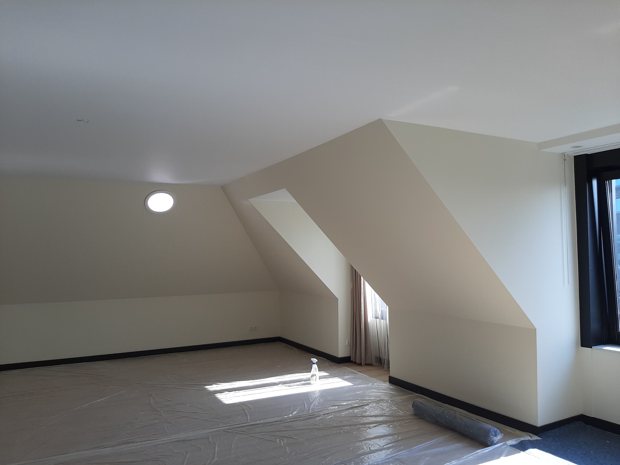 Jasne poddasze po odświeżeniu ścian, zabezpieczone folią malarską. Widoczne skosy, okrągłe okno i rolka materiału. Świeżo pomalowane ściany w neutralnym odcieniu.