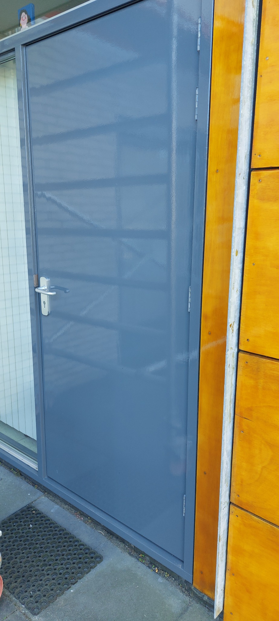 Szare, metalowe drzwi po malowaniu, z widocznymi zawiasami i klamką, przylegające do drewnianej ściany. Widoczny fragment wycieraczki przed drzwiami.