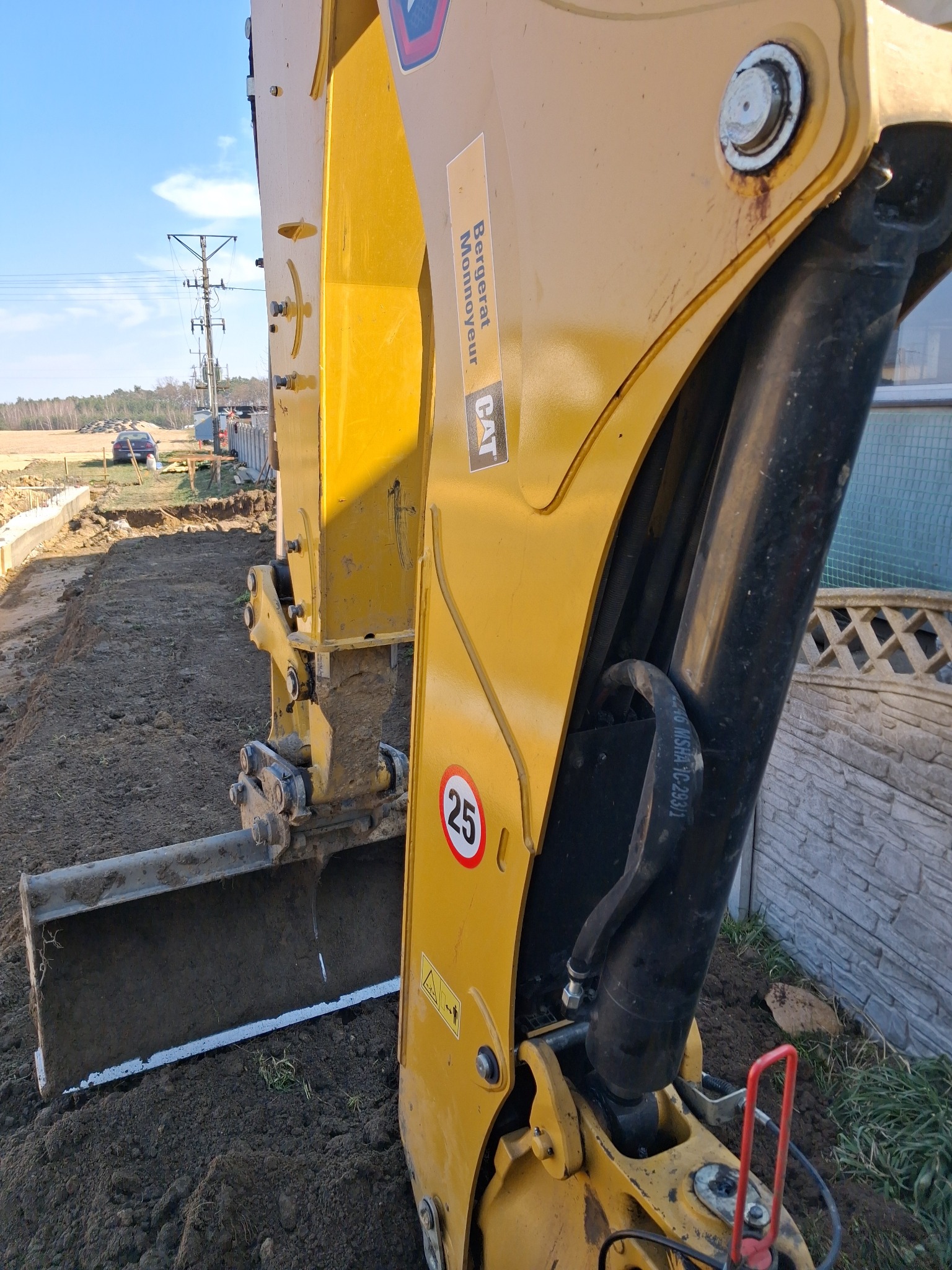 Żółta koparka CAT w trakcie prac ziemnych, widoczny wykop pod fundamenty i częściowo ogrodzenie. Na pierwszym planie łyżka koparki.