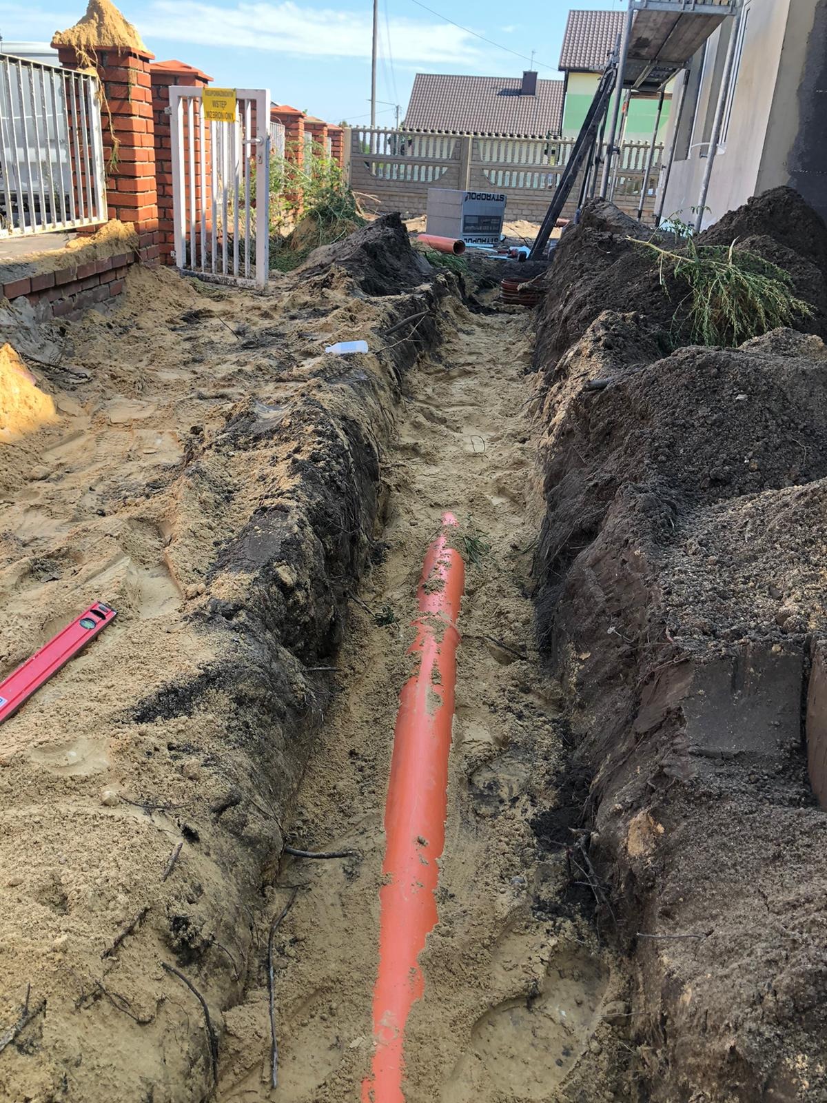 Wąski wykop w piaszczystej ziemi z pomarańczową rurą kanalizacyjną na dnie. W tle widoczny mur z cegły i brama. Po lewej poziomica.