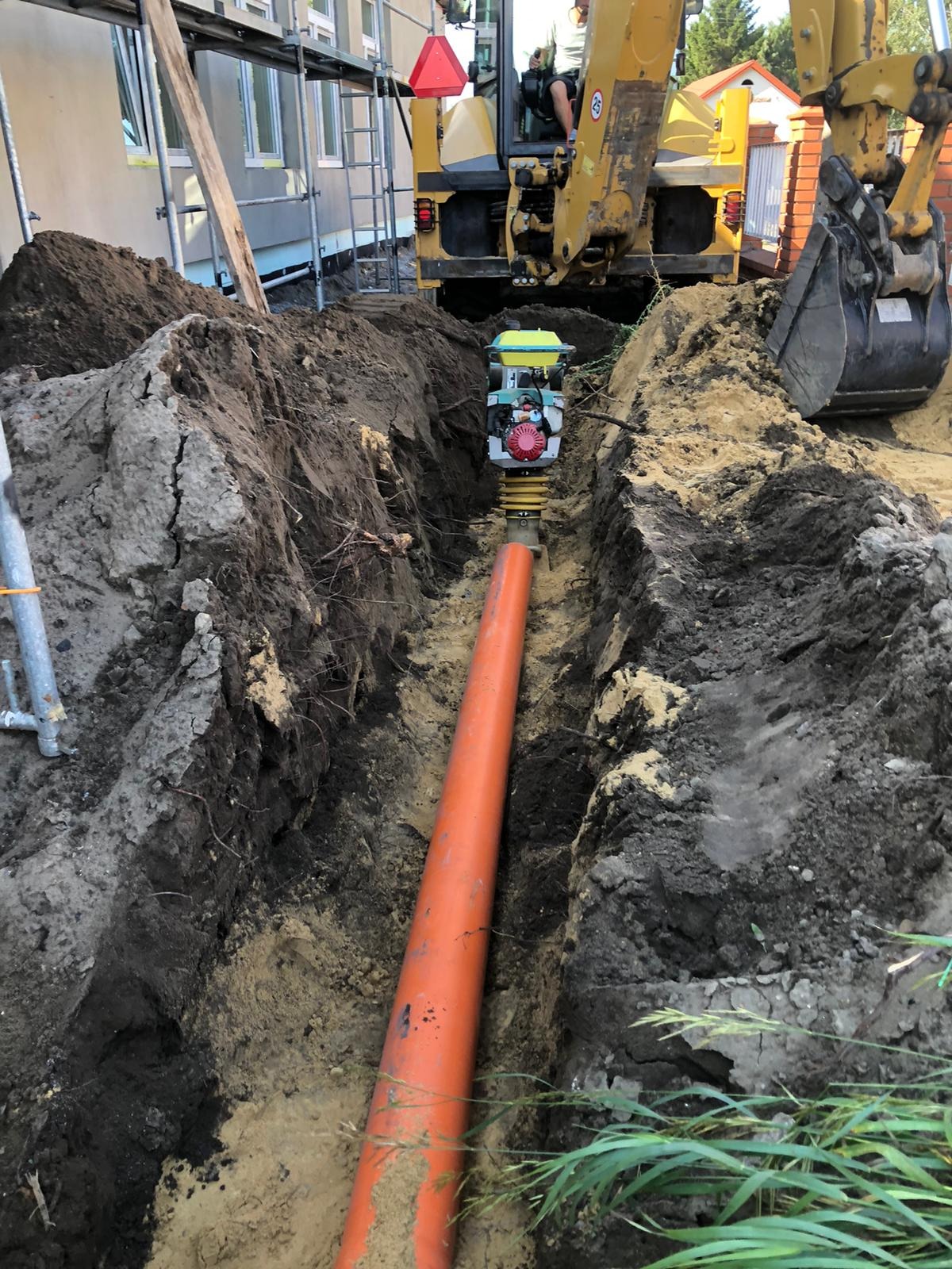 Wąski wykop z pomarańczową rurą i ubijarką wibracyjną, w tle koparka. Prace ziemne na budowie domu jednorodzinnego, widoczne rusztowania i fragment budynku.