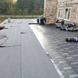 Volodymyr Tokar - Prace dekarskie na płaskim dachu: dekarz przy ścianie budynku, rolki papy Izobud, butle gazowe, narzędzia. Częściowo pokryty dach z widoczną membraną.