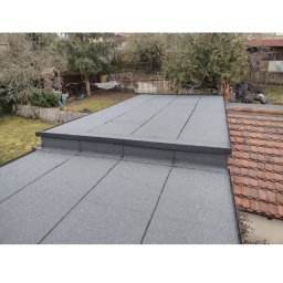 Volodymyr Tokar - Szary, płaski dach pokryty papą termozgrzewalną, widok z góry na tle zieleni i sąsiedniego dachu z czerwonej dachówki. Widoczne łączenia papy.