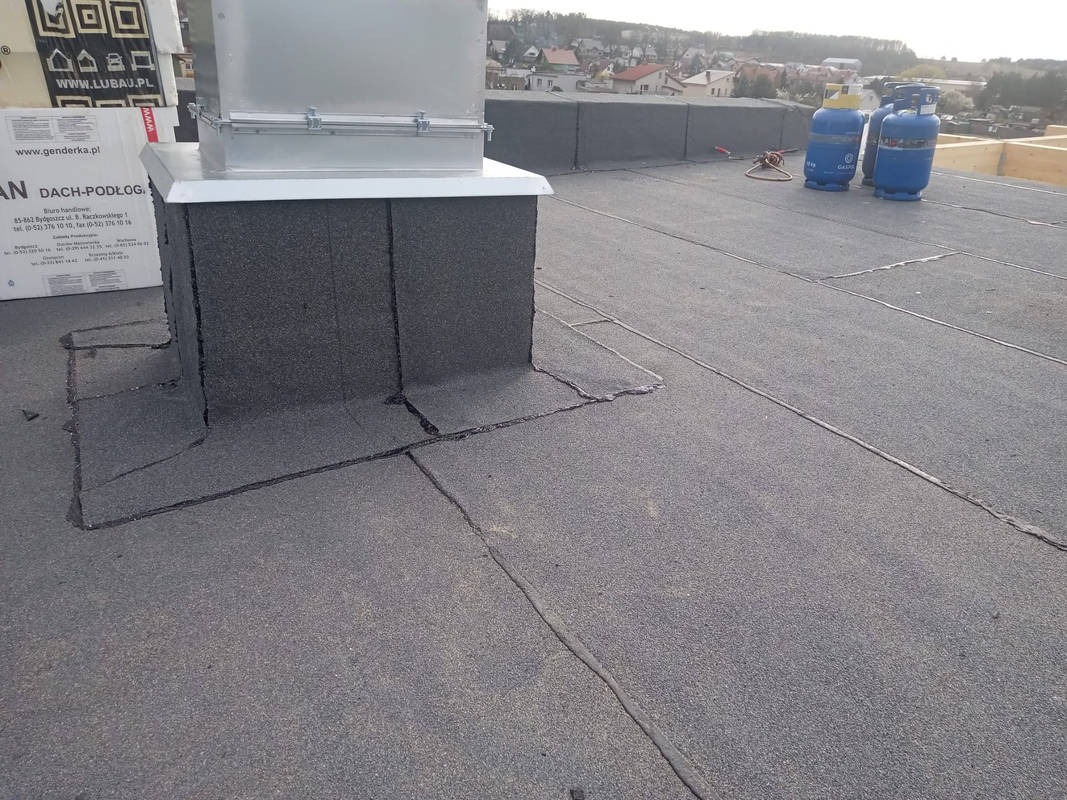 Wykończony dach płaski z papy termozgrzewalnej, z widoczną obróbką komina i butlami gazowymi w tle. Detal wykonania izolacji na dachu budynku.