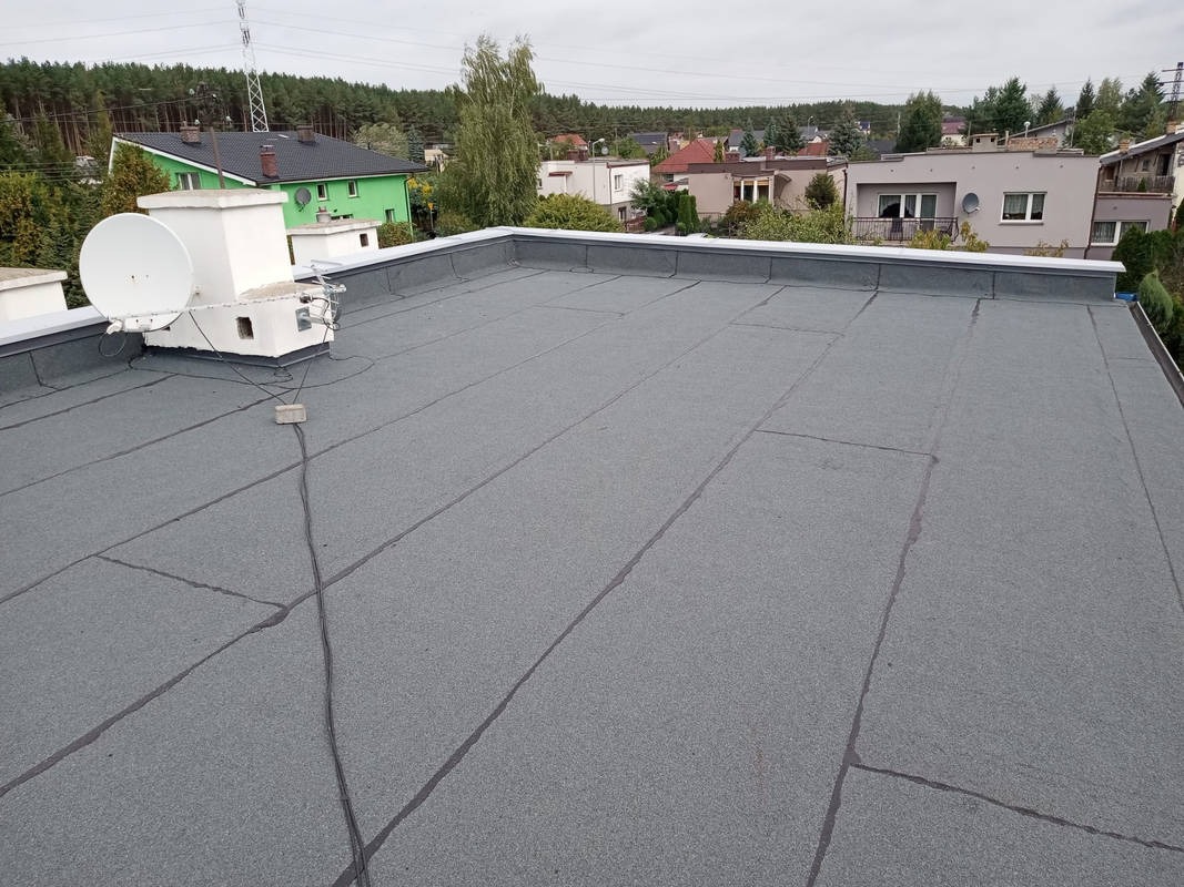 Szary, płaski dach pokryty papą termozgrzewalną, z widoczną anteną satelitarną i elementami obróbki blacharskiej. W tle zabudowa jednorodzinna i drzewa.