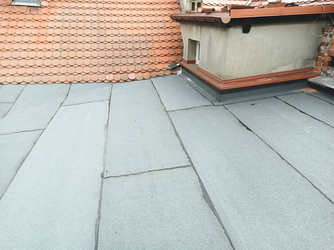 Szczegółowe ujęcie płaskiego dachu pokrytego papą termozgrzewalną we Wrocławiu, z widocznym fragmentem lukarny i dachu z dachówki karpiówki w tle.