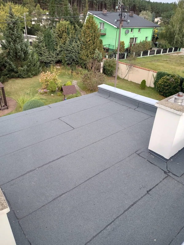 Szary, płaski dach z papy termozgrzewalnej na tle zielonego domu i drzew. Widoczne łączenia papy i komin. Ujęcie z góry, prezentujące wykończenie dachu.