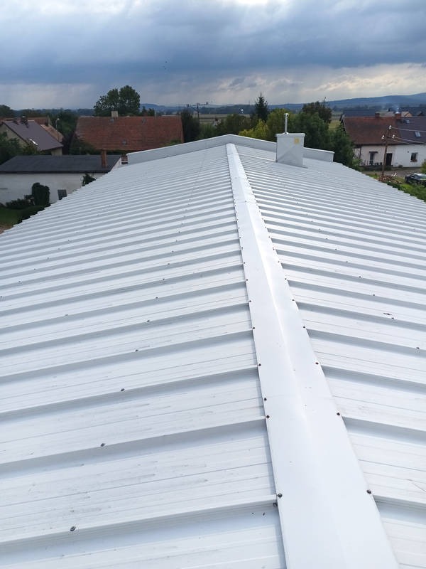 Biały, blaszany dach z widocznym kominem i elementami mocującymi, na tle zabudowań i pochmurnego nieba. Widok z góry.