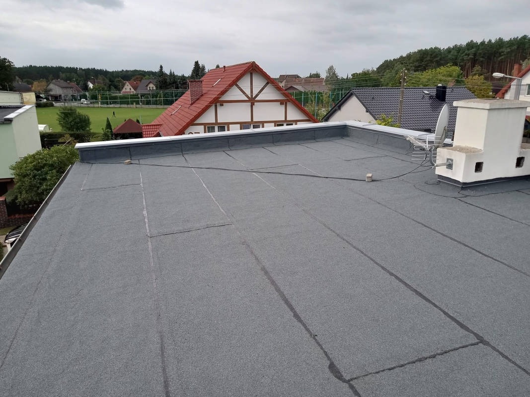 Szary, płaski dach pokryty papą termozgrzewalną z widocznym kominem i fragmentami zabudowy w tle, wykonany z dbałością o detale i estetykę.