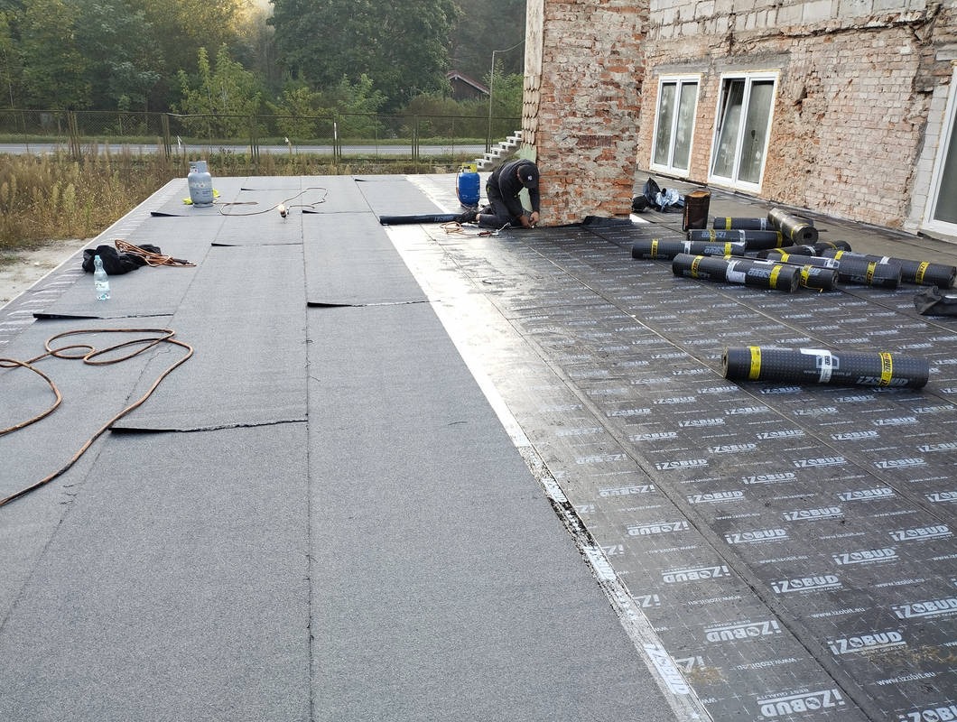 Prace dekarskie na płaskim dachu: dekarz przy ścianie budynku, rolki papy Izobud, butle gazowe, narzędzia. Częściowo pokryty dach z widoczną membraną.