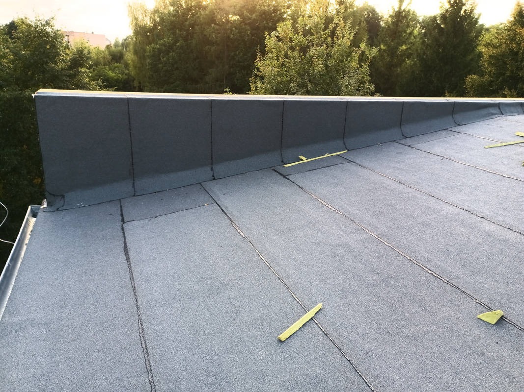 Szary dach płaski z papy termozgrzewalnej, widoczne łączenia i fragment obróbki blacharskiej. W tle zieleń drzew i fragment budynku. Detal dachu po renowacji.