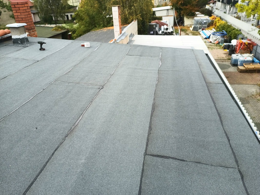 Szczelny, płaski dach pokryty papą termozgrzewalną z widocznym kominem, narzędziem i elementami wentylacji. Tło stanowią budynki i drzewa.