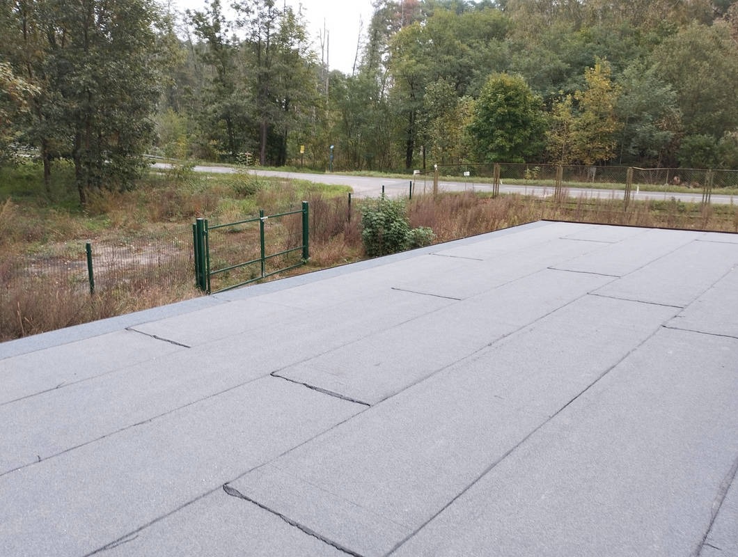 Szary, płaski dach pokryty papą termozgrzewalną, widok z góry. W tle zieleń drzew i krzewów oraz ogrodzenie z siatki.