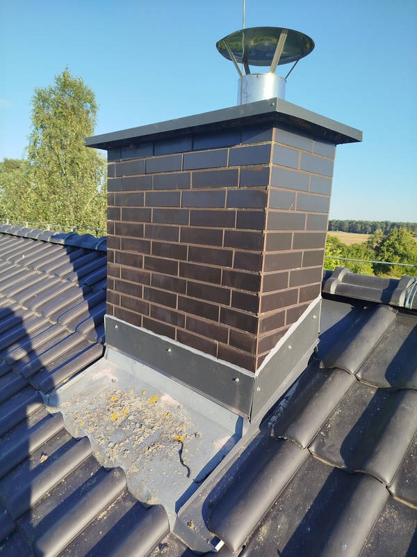 Ciemna, ceglana obudowa komina z metalowym daszkiem na dachu krytym ciemną dachówką. Widoczne obróbki blacharskie i zanieczyszczenia na dachu. Ujęcie z góry.