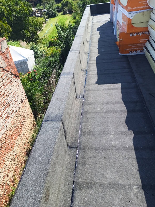 Wykończony, płaski dach pokryty papą z widocznymi obróbkami blacharskimi i materiałami izolacyjnymi Baudepir. Ujęcie z góry, w tle zieleń i budynek.