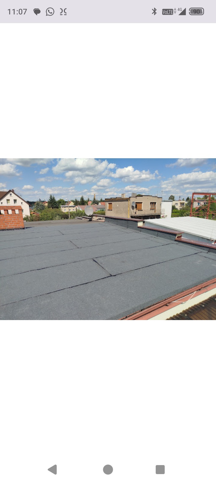 Płaski dach pokryty papą termozgrzewalną na budynku z ceglanym kominem. W tle zabudowa miejska i błękitne niebo z chmurami.