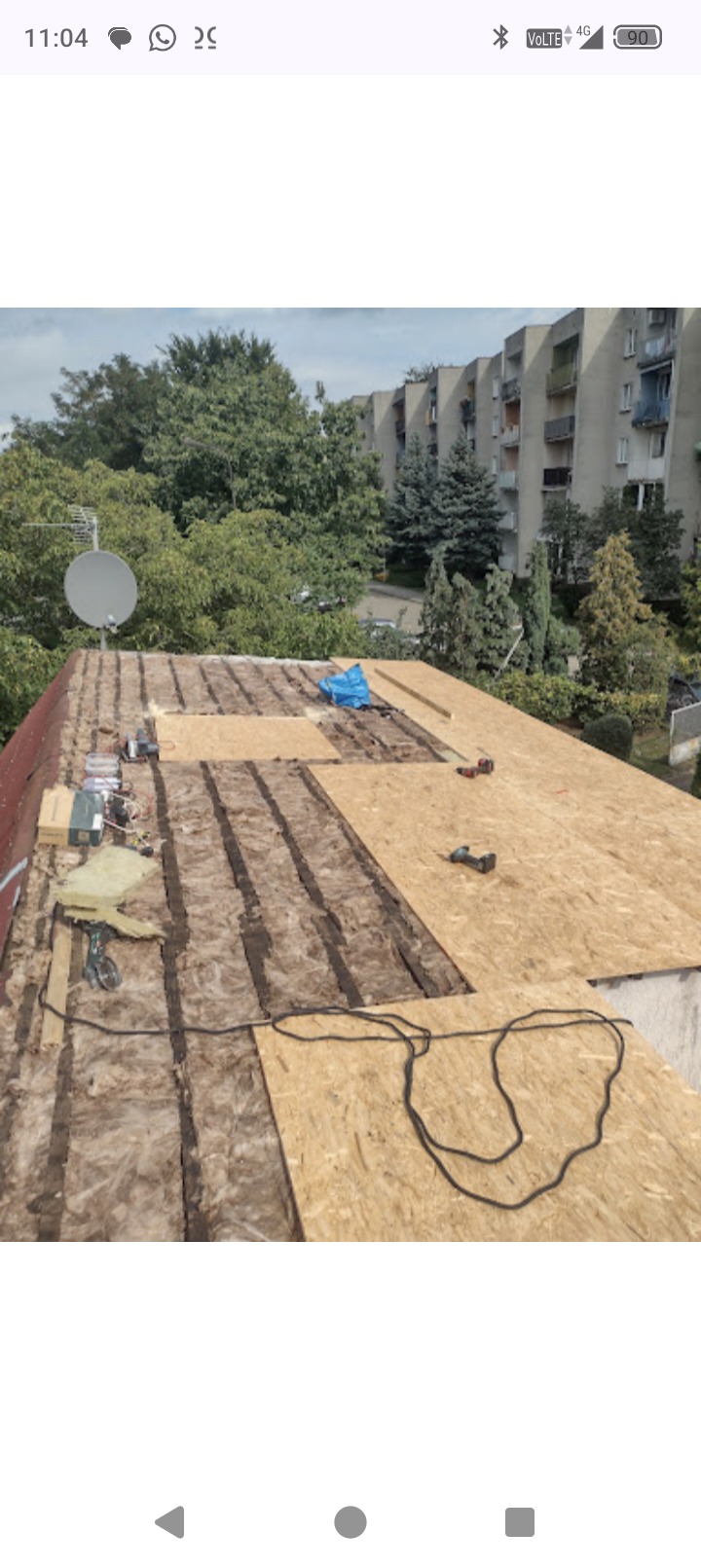 Dach w trakcie remontu: widoczne warstwy izolacji, płyty OSB, narzędzia. W tle budynek mieszkalny i zieleń. Ujęcie z góry, ukazujące postęp prac.