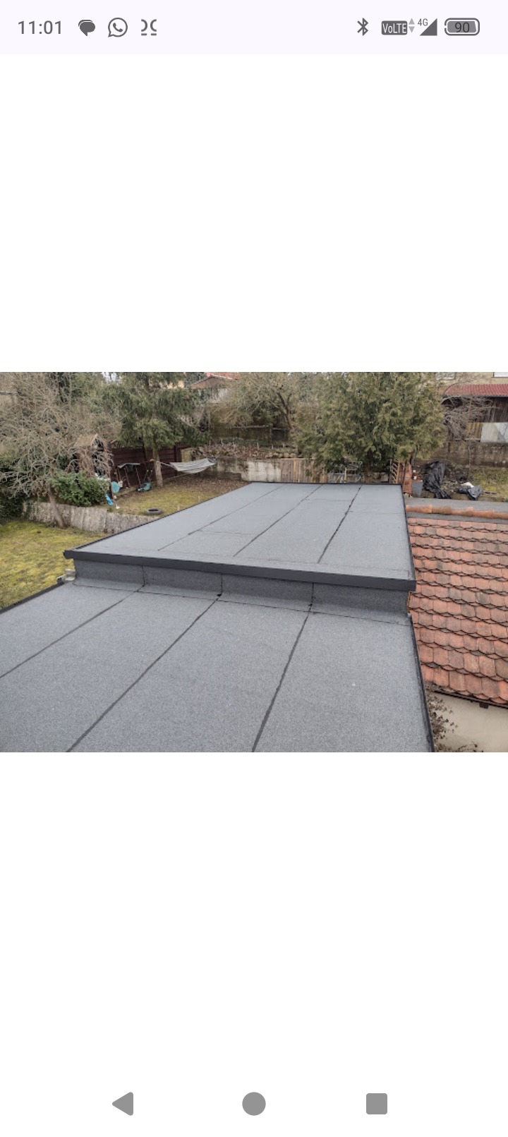 Szary, płaski dach pokryty papą termozgrzewalną, widok z góry na tle zieleni i sąsiedniego dachu z czerwonej dachówki. Widoczne łączenia papy.