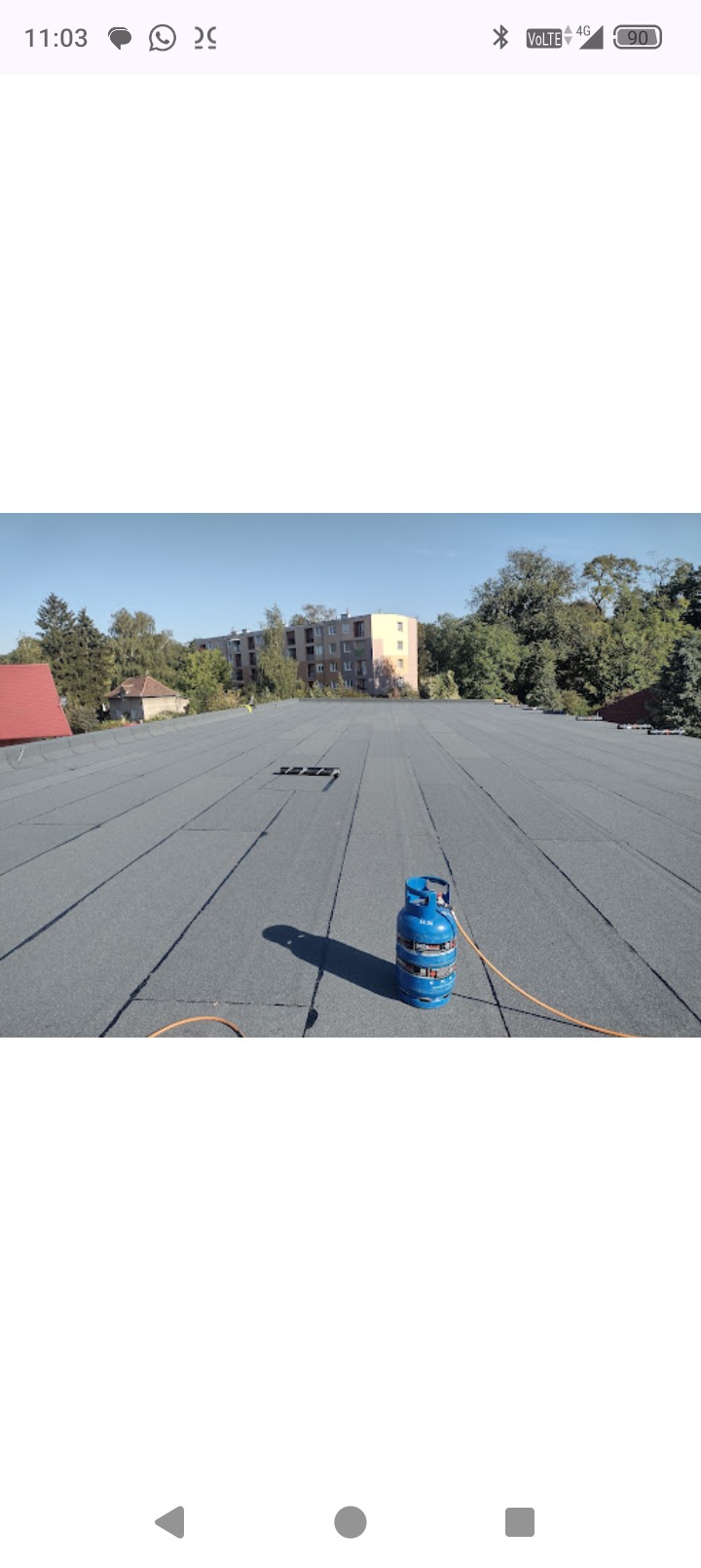 Szary dach pokryty papą termozgrzewalną z widocznym butlą gazową i budynkiem w tle. Układanie papy na dachu.