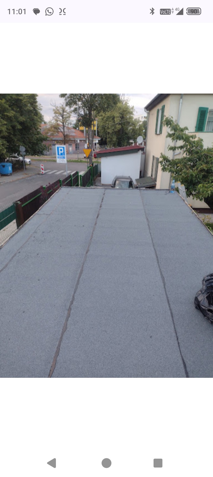 Szary, płaski dach pokryty papą termozgrzewalną, widok z góry. W tle budynki i drzewa. Na dachu leżą buty robocze.