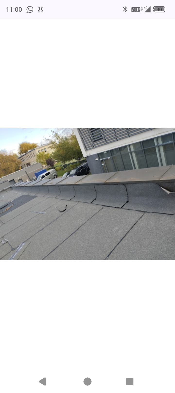 Uszkodzony, płaski dach pokryty papą na tle budynku biurowego i drzew. Widoczne pęknięcia i nierówności na powierzchni dachu. Ujęcie z bliska, podkreślające stan techniczny.