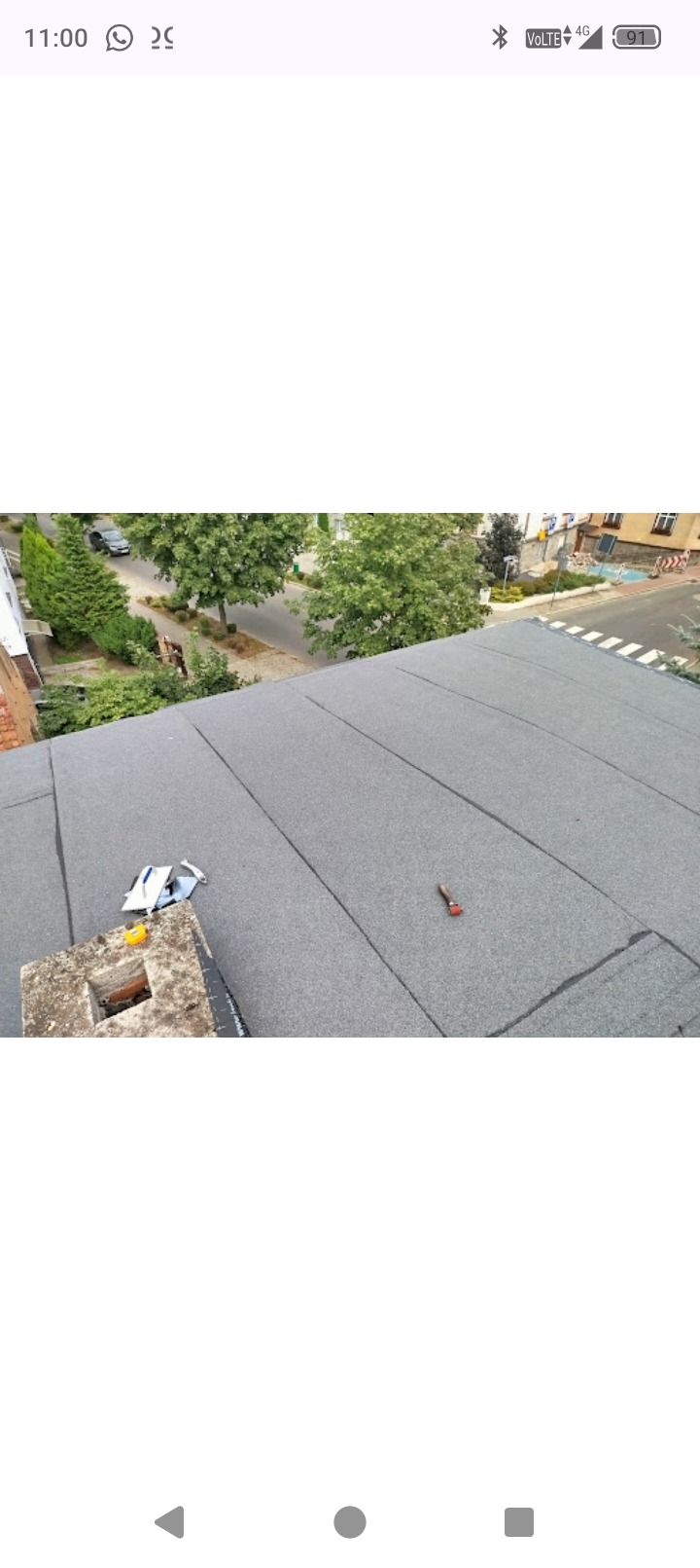 Szary, płaski dach pokryty papą termozgrzewalną, widok z góry. Na dachu narzędzia dekarskie i komin. W tle zieleń drzew i zabudowa miejska.