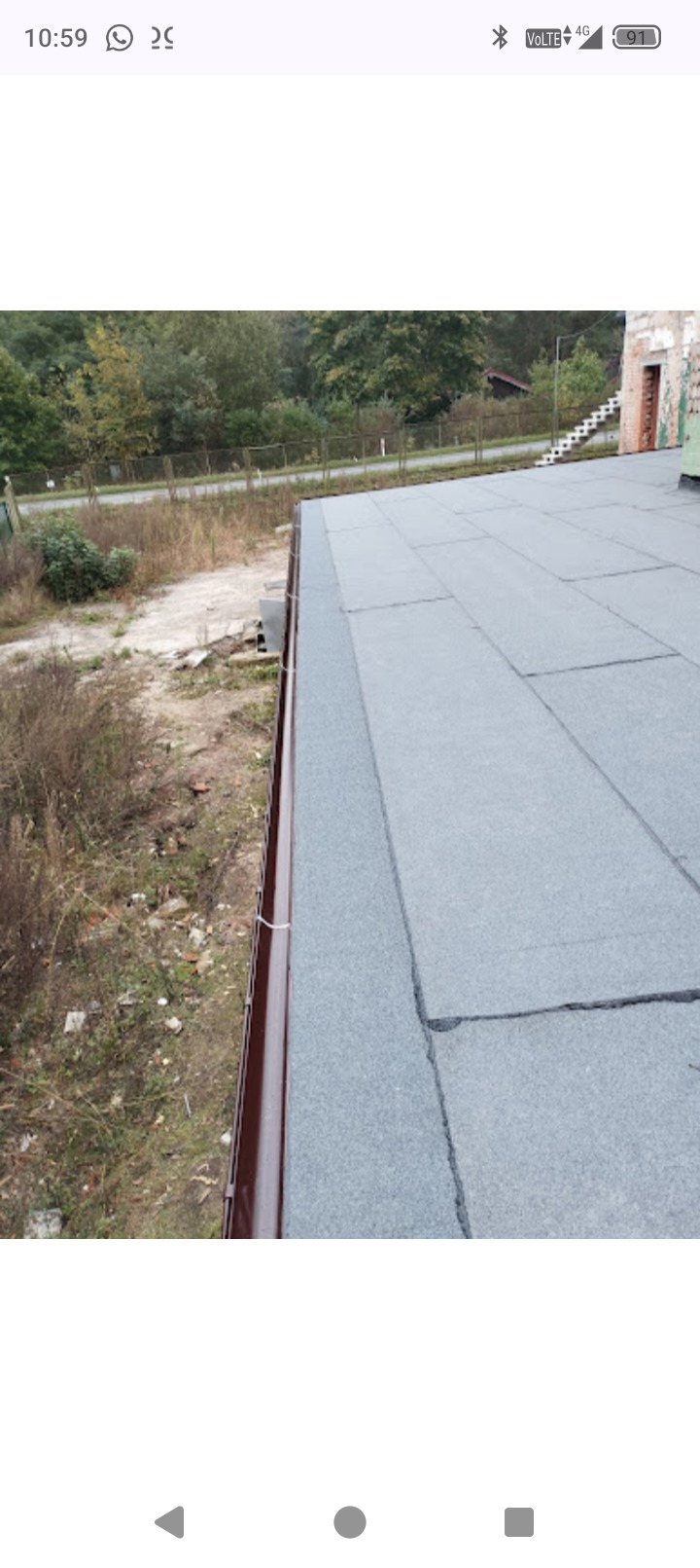 Szary dach pokryty papą z brązową rynną wzdłuż krawędzi. Widok z góry na budynek i otaczającą go zieleń. Detal wykonania pokrycia dachowego.