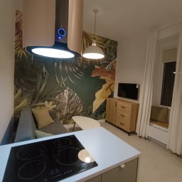 K1 - REMONTY & DOCIEPLENIA PRZEMYSŁAW KULKA - Nowoczesne wnętrze: płyta indukcyjna na wyspie kuchennej, tropikalna tapeta, szara sofa z poduszkami, drewniana komoda i okno z siedziskiem. Całość w ciepłej tonacji.