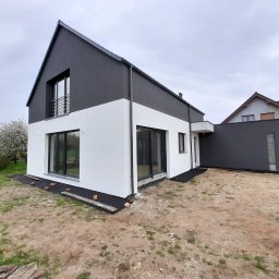 K1 - REMONTY & DOCIEPLENIA PRZEMYSŁAW KULKA - Nowoczesny dom jednorodzinny z biało-grafitową elewacją, duże okna, widok z zewnątrz, teren wokół domu w trakcie zagospodarowywania, pochmurne niebo.