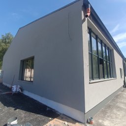 K1 - REMONTY & DOCIEPLENIA PRZEMYSŁAW KULKA - Szary budynek po otynkowaniu z czarnymi oknami i rynną, widok z zewnątrz w słoneczny dzień. Na pierwszym planie narzędzia i materiały budowlane.