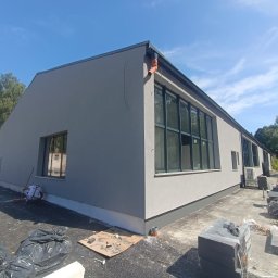 K1 - REMONTY & DOCIEPLENIA PRZEMYSŁAW KULKA - Szary budynek po ociepleniu elewacji z czarnymi oknami. Pracownik zamiata teren wokół budynku. Widoczne materiały budowlane i narzędzia.