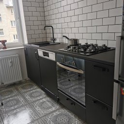 K1 - REMONTY & DOCIEPLENIA PRZEMYSŁAW KULKA - Ciemna zabudowa kuchenna z białymi płytkami na ścianie i wzorzystą podłogą. Na blacie kuchenka gazowa z garnkiem. Widok na okno z grzejnikiem i narzędziem.