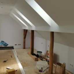 Białe poddasze ze skośnymi oknami dachowymi, drewnianymi belkami i narzędziami malarskimi. Widoczne przygotowania do malowania ścian.