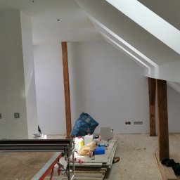 K1 - REMONTY & DOCIEPLENIA PRZEMYSŁAW KULKA - Białe, świeżo pomalowane poddasze z drewnianymi belkami, widoczne narzędzia i materiały budowlane, okno dachowe wpuszczające światło. Wnętrze w trakcie wykańczania.
