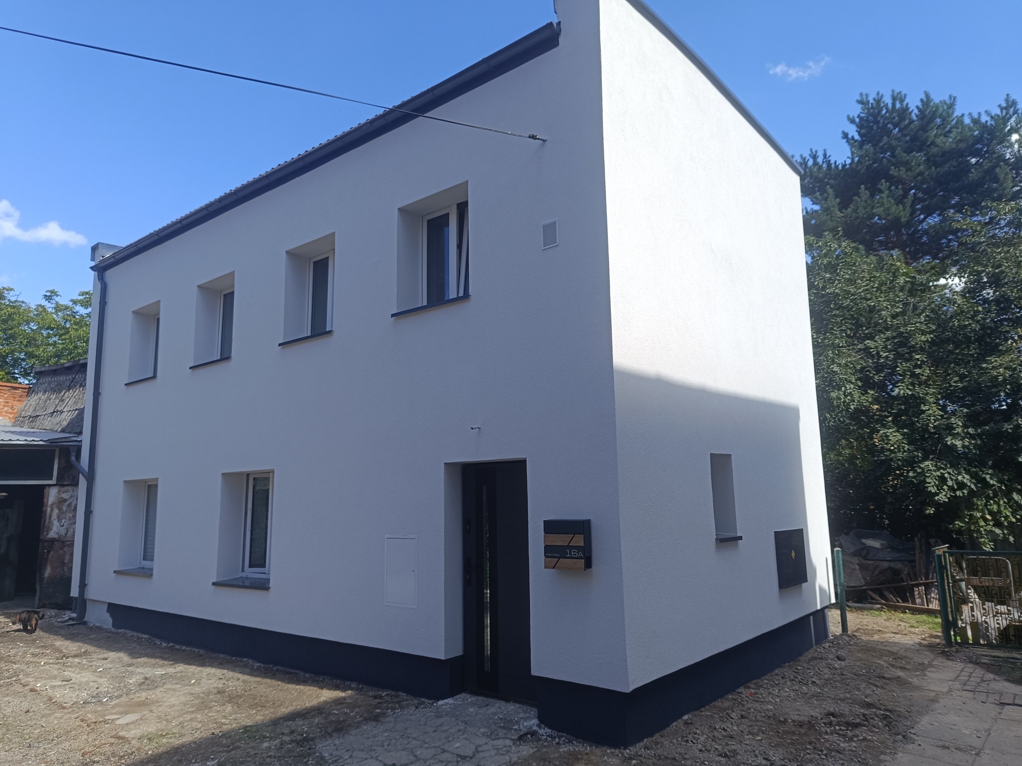 Odnowiona elewacja dwukondygnacyjnego domu z nową, białą fasadą i ciemnymi akcentami przy oknach i podstawie. Widoczna skrzynka pocztowa z numerem 16A.