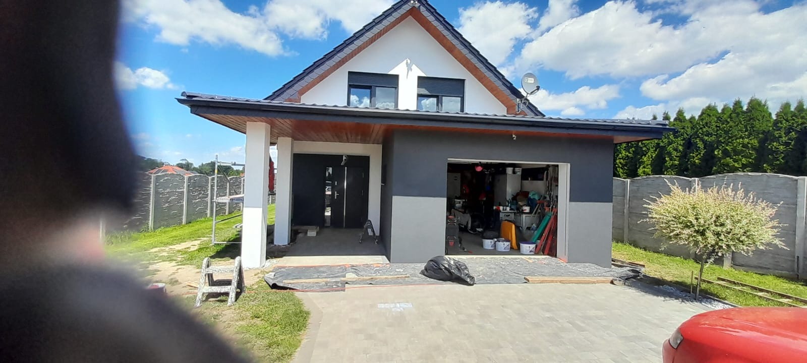 Dwukondygnacyjny dom z garażem w trakcie malowania elewacji na szaro, z widocznymi narzędziami i zabezpieczeniami. Częściowo zasłonięty kadr, słoneczny dzień.