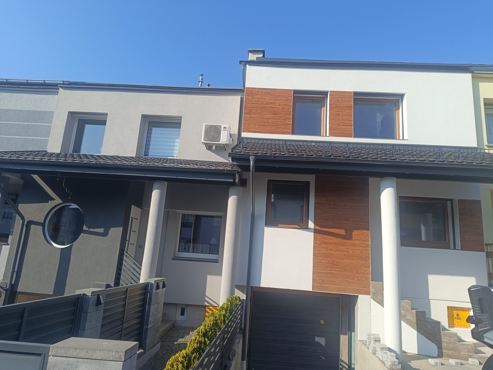 Elewacja szeregowca z elementami drewnopodobnymi i klimatyzacją. Szara i biała kolorystyka, nowoczesny design. Widoczne schody wejściowe i fragment ogrodzenia.