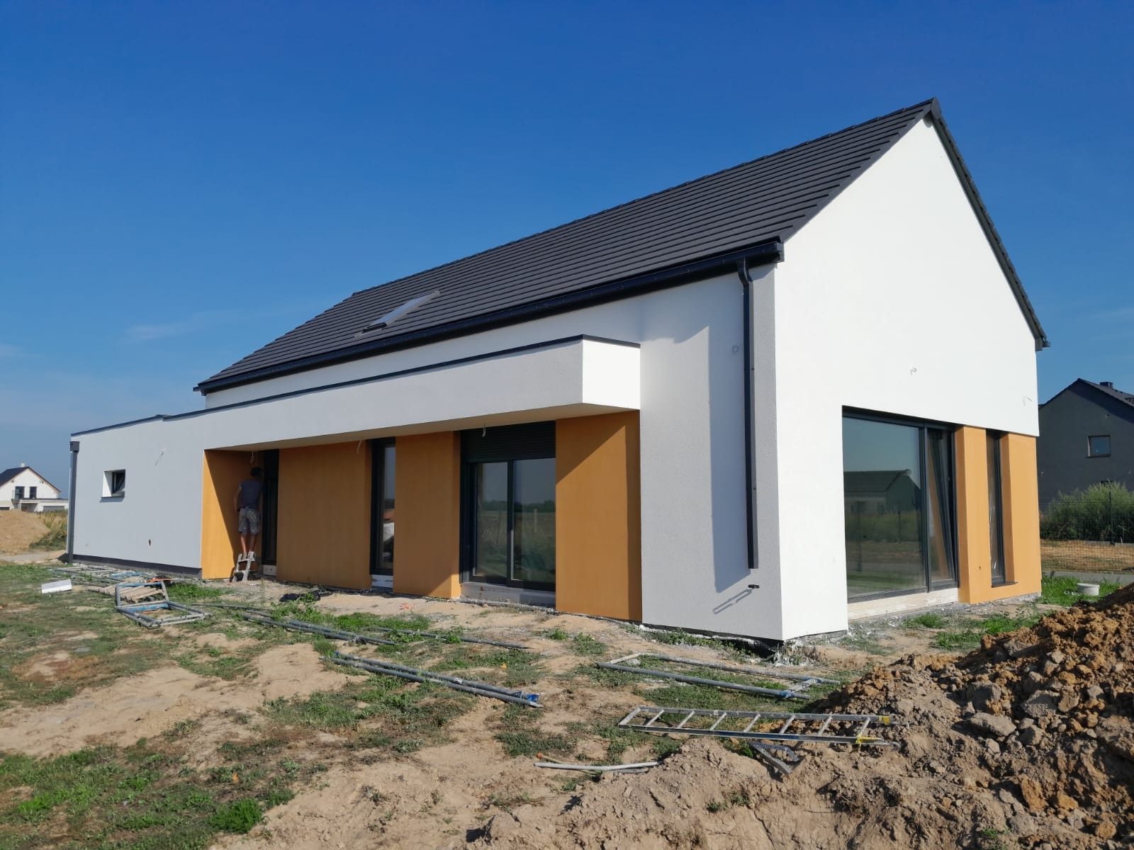 Nowoczesny dom jednorodzinny z białymi ścianami i elementami w kolorze ochry, w trakcie prac wykończeniowych. Na pierwszym planie drabina i rury.