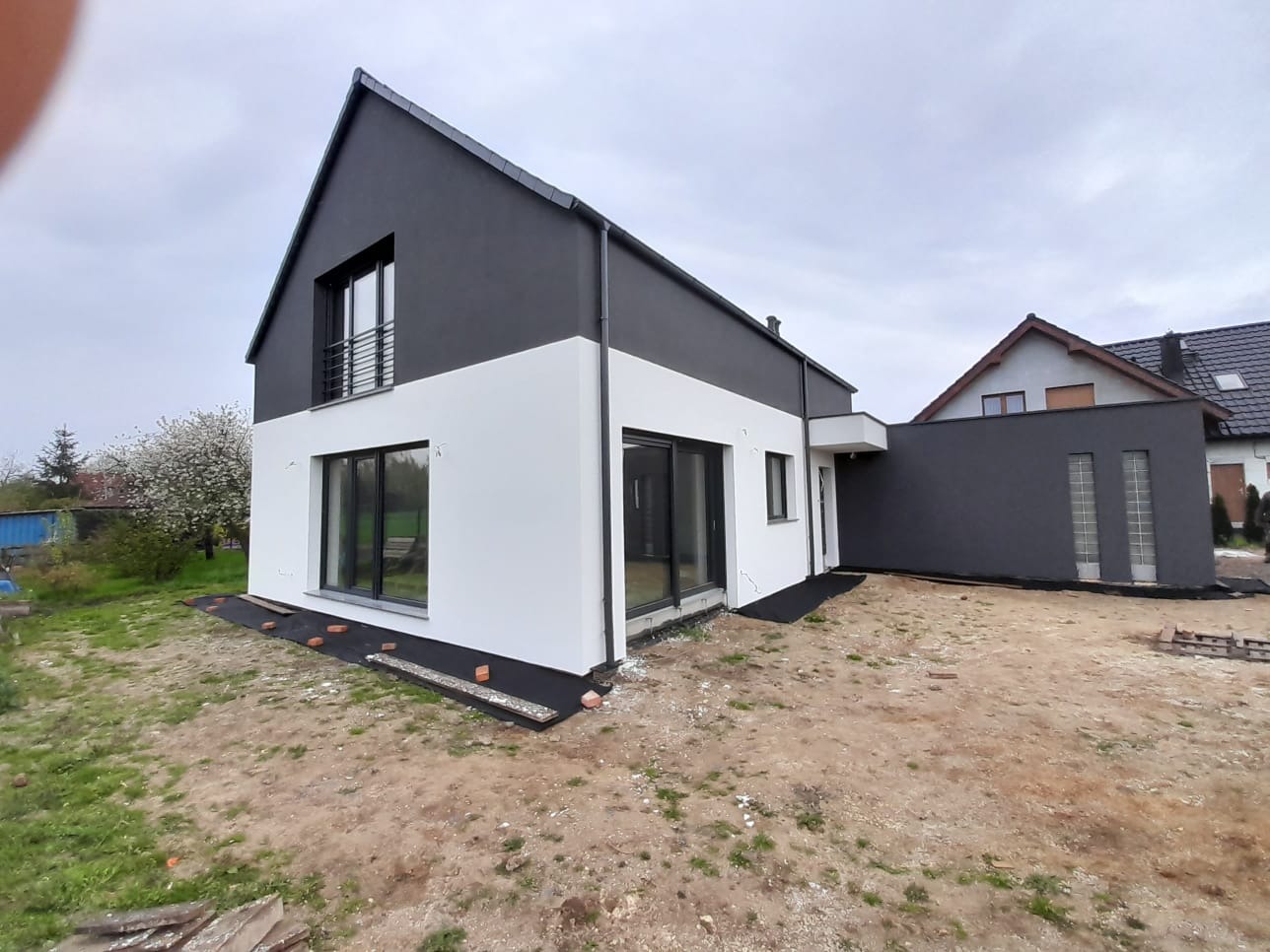 Nowoczesny dom jednorodzinny z biało-grafitową elewacją, duże okna, widok z zewnątrz, teren wokół domu w trakcie zagospodarowywania, pochmurne niebo.