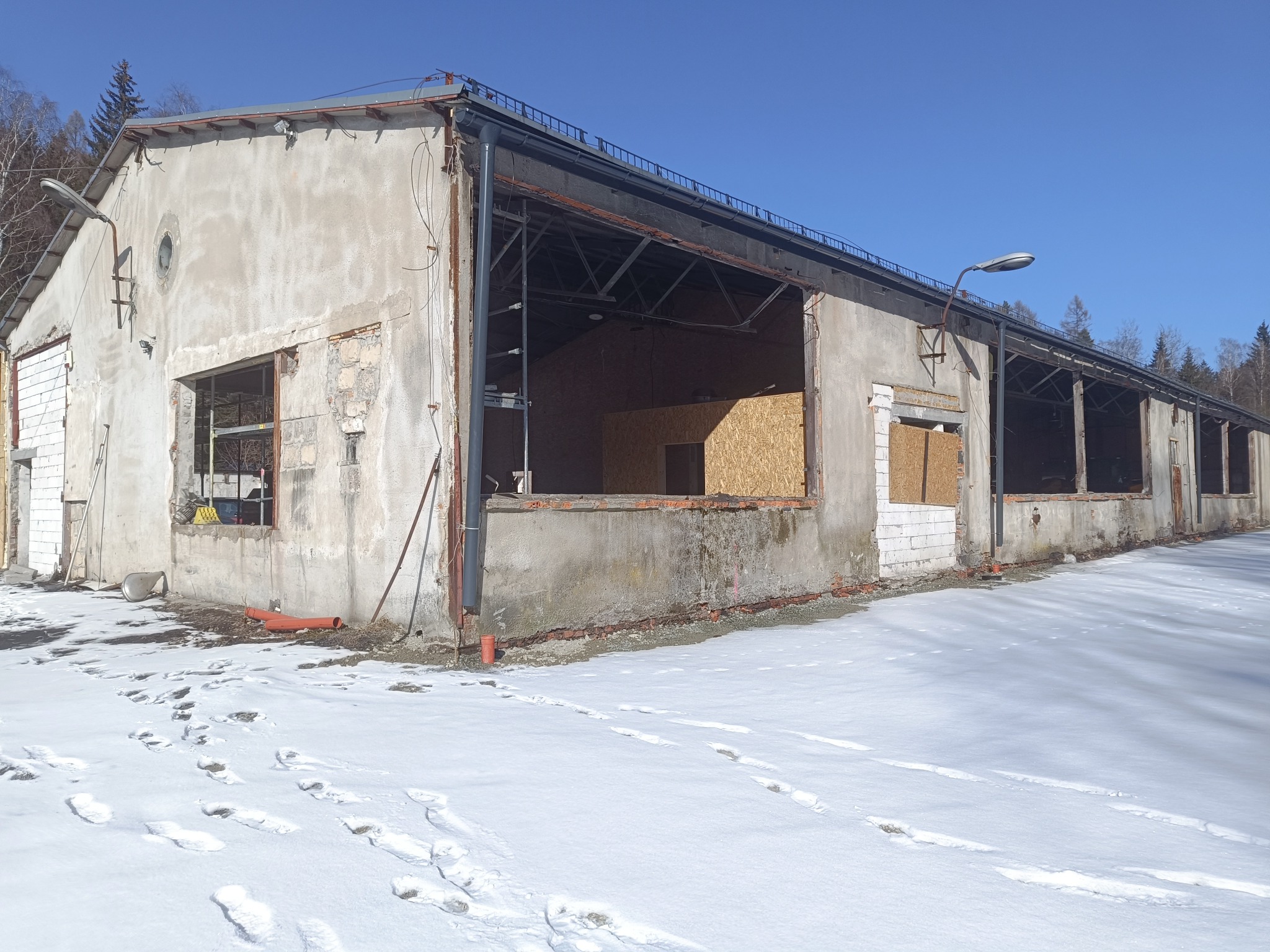 Budynek w trakcie remontu z częściowo zamurowanymi oknami i śladami na śniegu. Widoczne elementy konstrukcyjne i materiały budowlane. Słoneczny dzień.