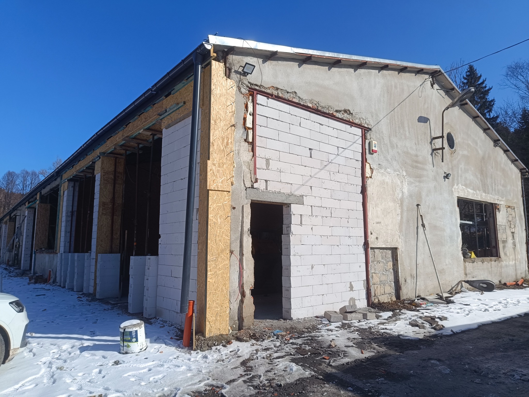 Budynek w trakcie ocieplania: fragment ściany z białych bloczków, obok płyty OSB. Widoczny śnieg i wiadro z farbą. Błękitne niebo w tle.