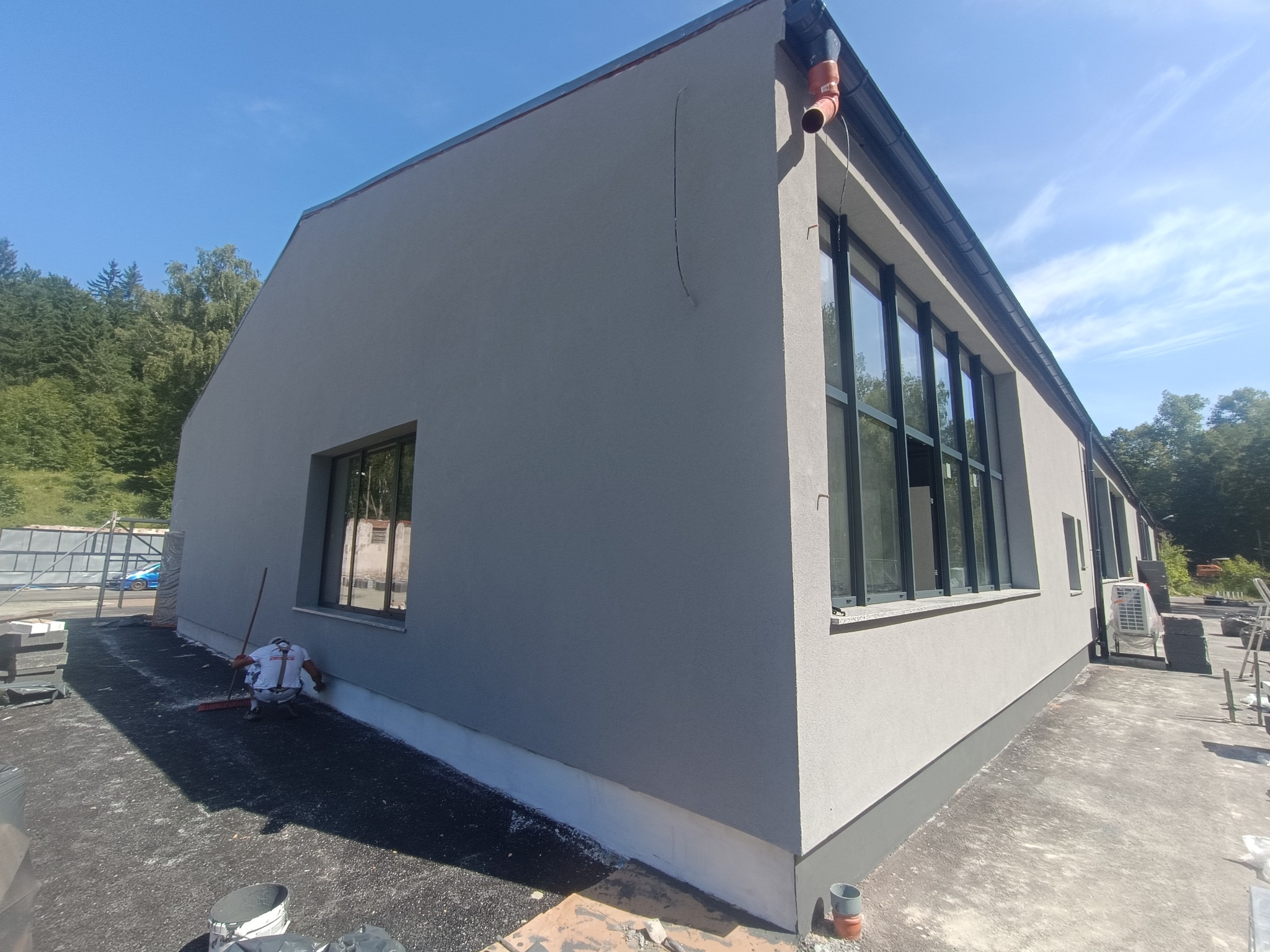 Szary budynek po otynkowaniu z czarnymi oknami i rynną, widok z zewnątrz w słoneczny dzień. Na pierwszym planie narzędzia i materiały budowlane.