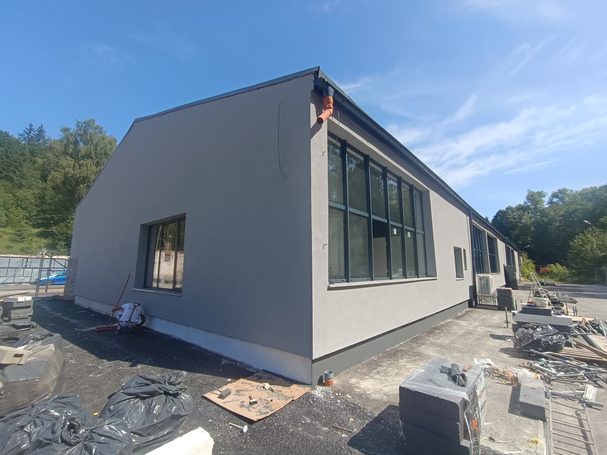 Szary budynek po ociepleniu elewacji z czarnymi oknami. Pracownik zamiata teren wokół budynku. Widoczne materiały budowlane i narzędzia.