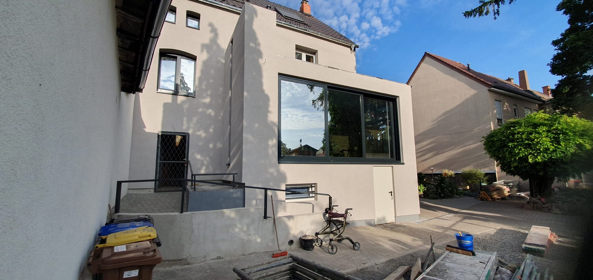 Elewacja domu po remoncie z dużym oknem i nowym wejściem. Widoczne śmietniki, rusztowanie i narzędzia malarskie. Jasna kolorystyka budynku i błękitne niebo w tle.