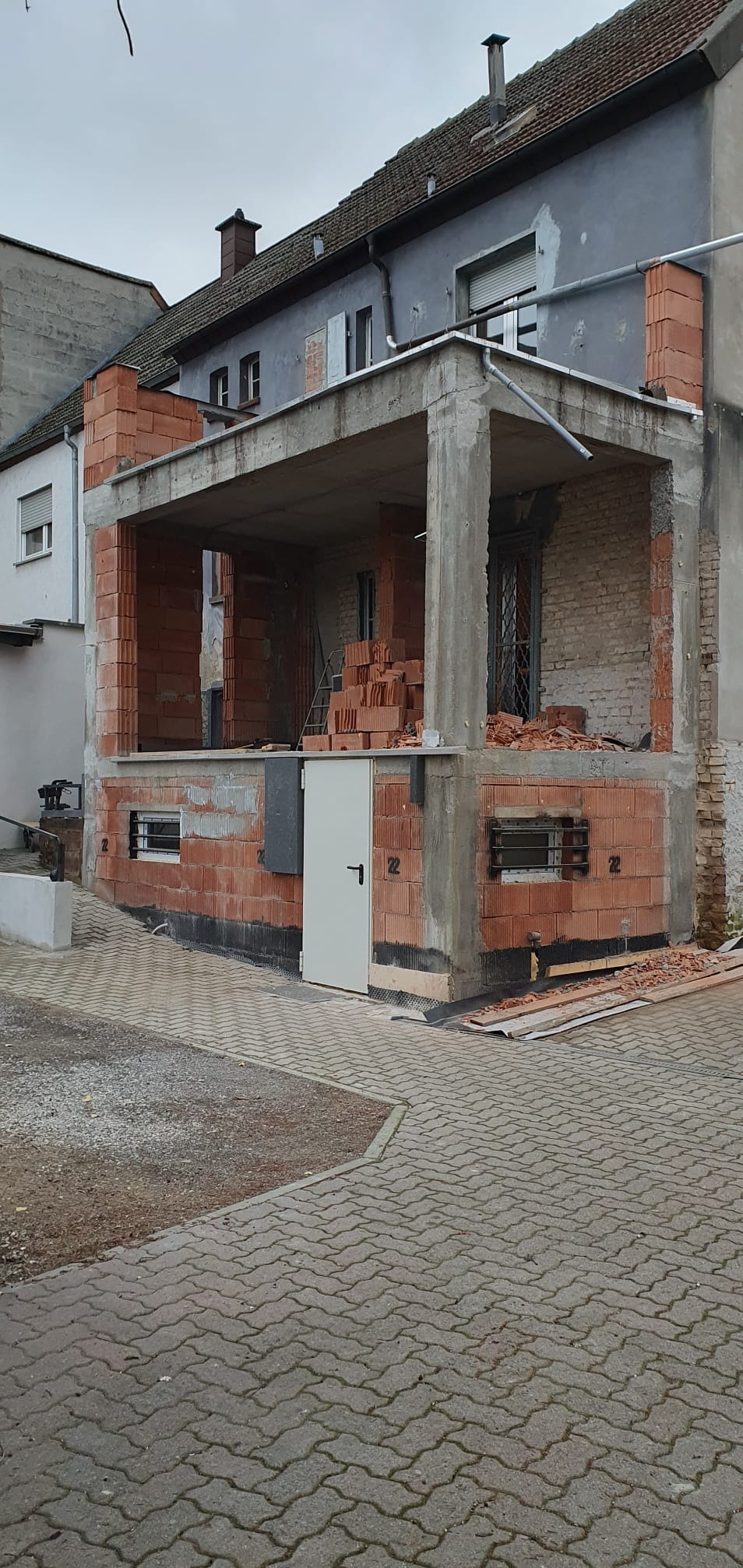 Budowa domu z czerwonej cegły: wznoszenie ścian i filarów, widoczne materiały budowlane, brama wejściowa. Prace murarskie w toku, surowy stan.