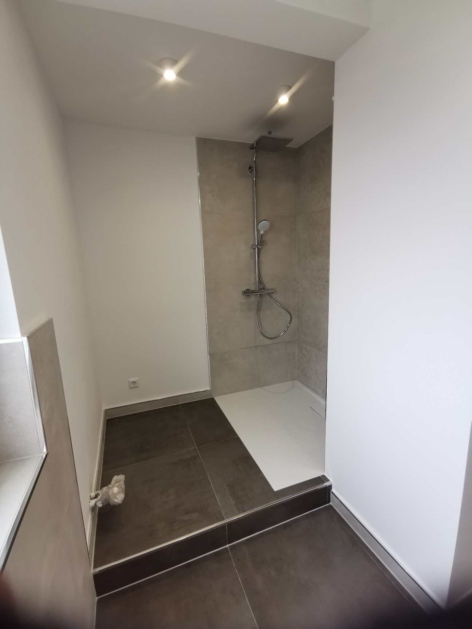 Nowoczesna kabina prysznicowa walk-in z szarymi płytkami i ciemną podłogą. Minimalistyczny design, oświetlenie punktowe. Widok na brodzik i baterię.