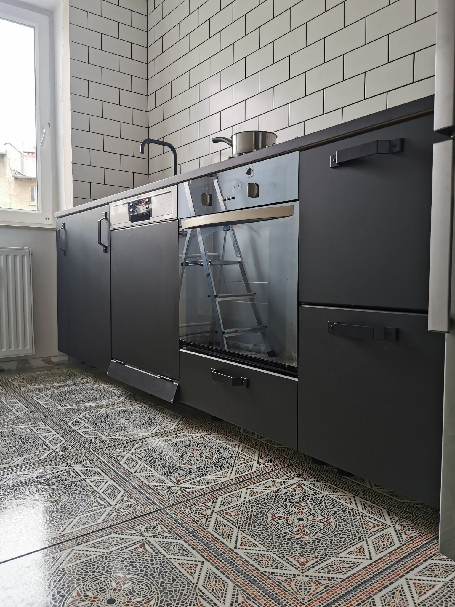 Nowoczesna kuchnia z czarnymi szafkami, białymi kafelkami na ścianie i wzorzystą podłogą. W piekarniku widoczna drabinka. Minimalistyczny design.