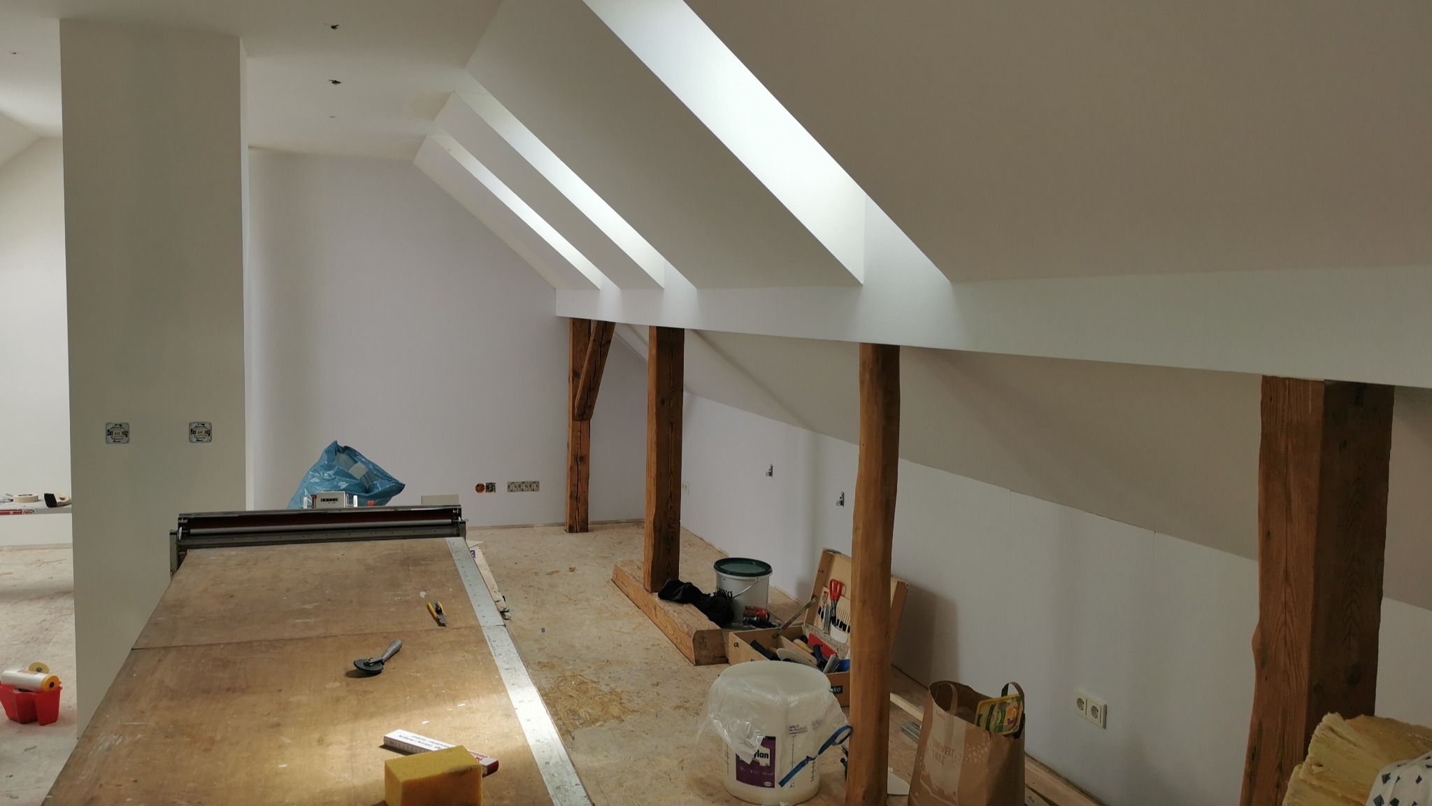 Białe poddasze ze skośnymi oknami dachowymi, drewnianymi belkami i narzędziami malarskimi. Widoczne przygotowania do malowania ścian.